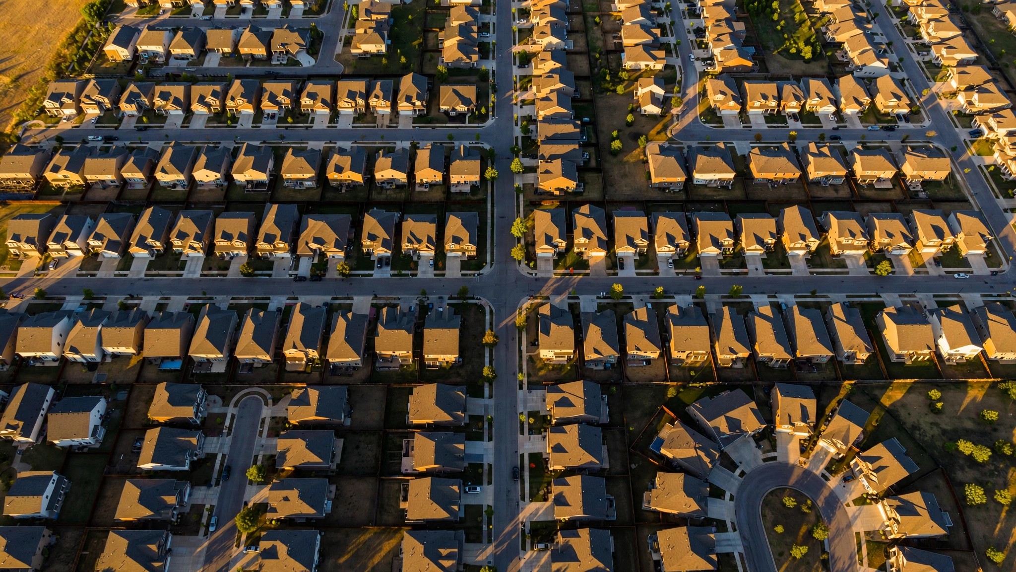 Aerial view of a production housing subdivision with identical homes arranged along curving streets, shadows falling at different angles across each lot