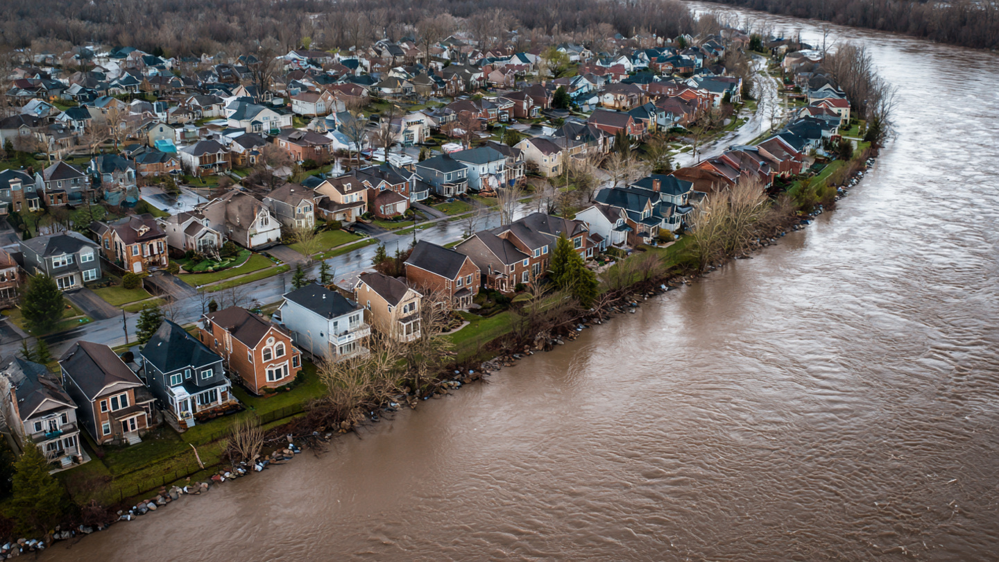 Aerial view of a suburban neighborhood next to a swollen river, overcast sky, houses near the waterline with sandbags visible