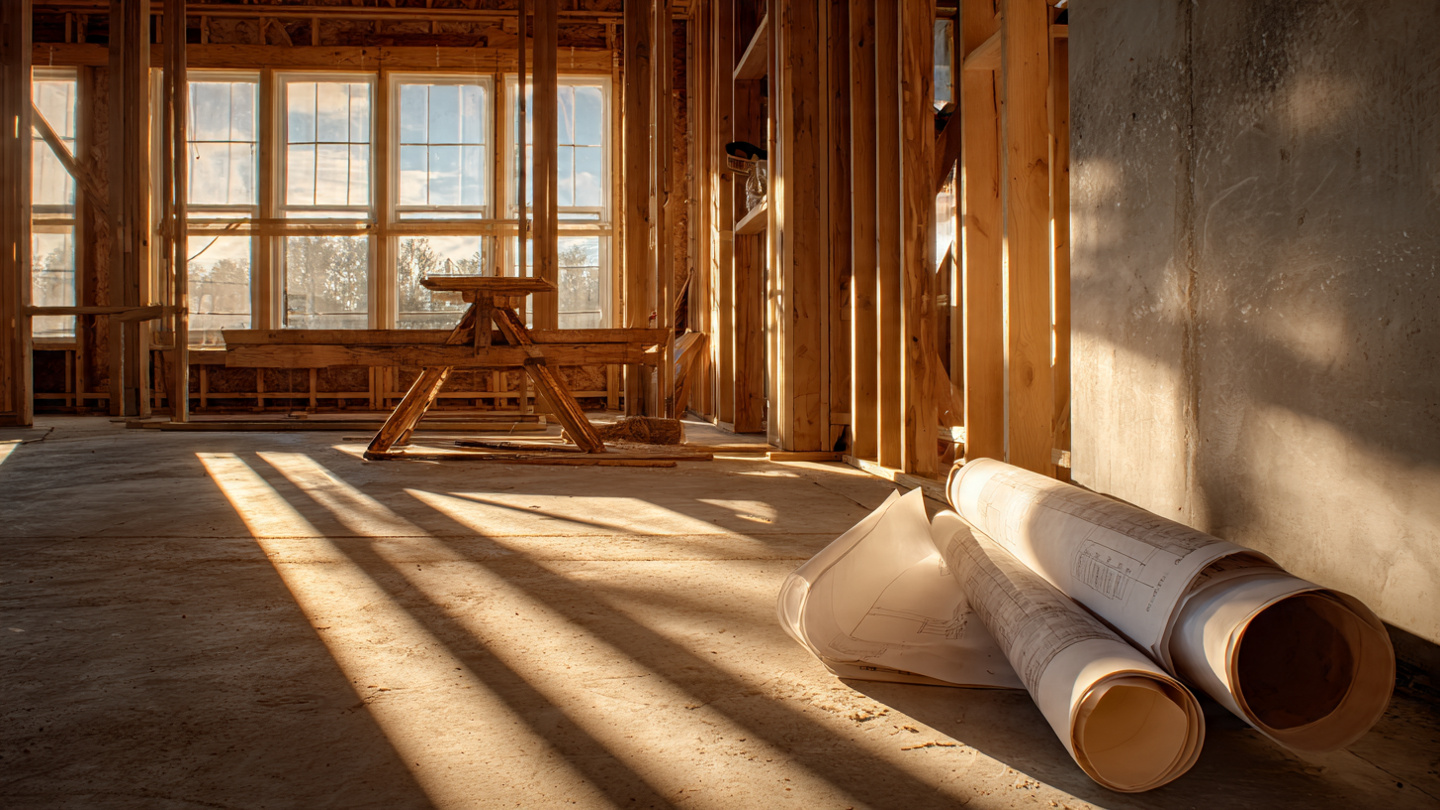 Sunlight streaming through tall windows of an unfinished residential home, casting long diagonal shadows across bare concrete floors, with architectural blueprints scattered on a sawhorse table