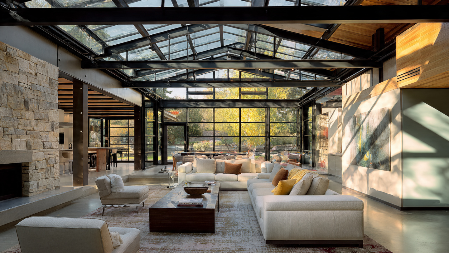 A modern residential living room with a dramatic floor-to-ceiling glass wall and exposed steel I-beams in the ceiling