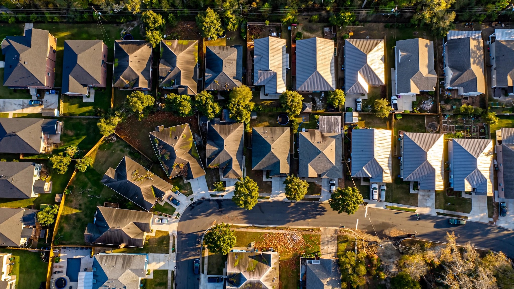 Aerial view of a residential neighborhood with rooftops of varying ages and conditions, late afternoon light casting long shadows across shingle patterns