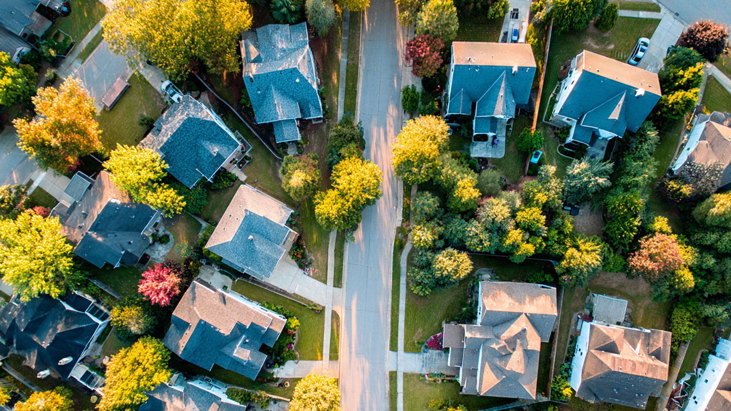 A residential roof with visible hail damage on asphalt shingles, pockmarked with dark bruise marks, with a clear blue sky and suburban neighborhood in the background
