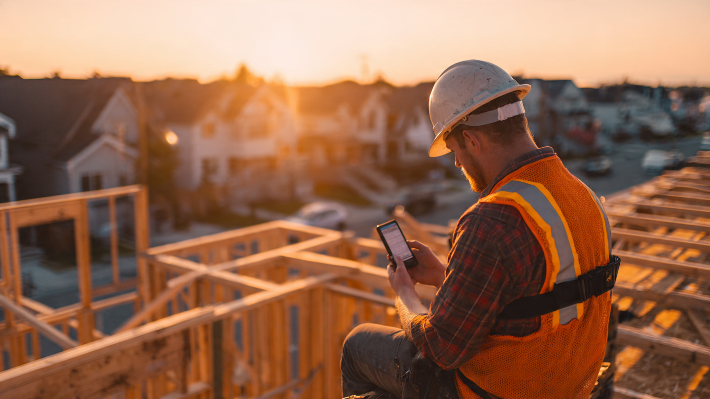 A construction worker on a residential roof framing job, a small GPS tracking device clipped to his tool belt, security cameras visible on a nearby pole, overcast morning light