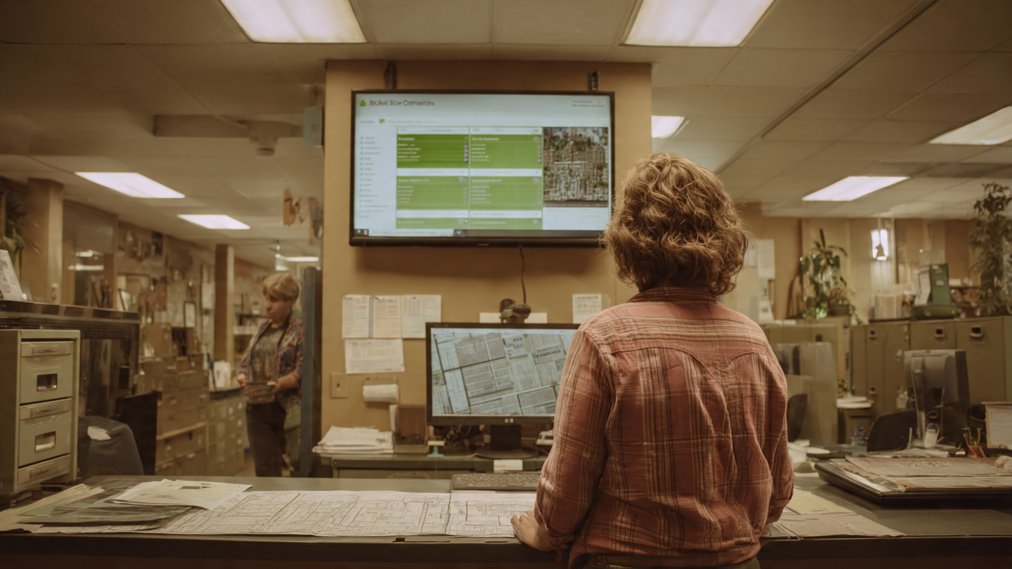 A homeowner at a city planning counter reviewing blueprints while a computer screen shows an AI zoning compliance interface