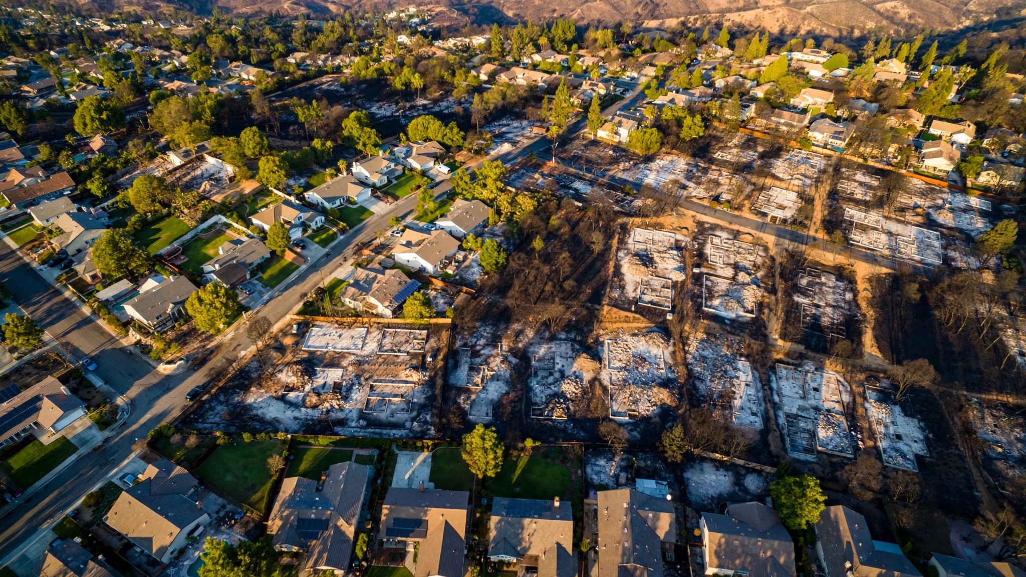 Aerial view of a wildfire-damaged neighborhood where some homes remain standing amid destroyed lots, showing the patchwork pattern of structure survival