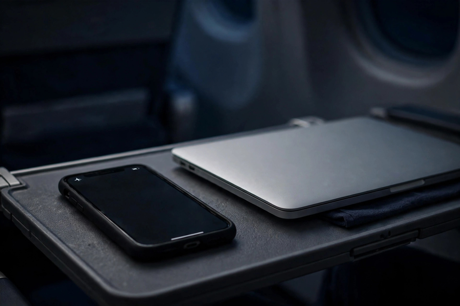 A phone in airplane mode on an airplane tray table beside a closed laptop, cabin light low