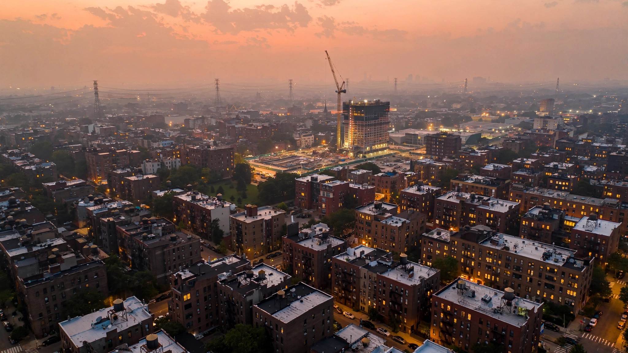 Overhead view of a New York City residential block at dusk, half the windows dark from a power outage while construction cranes for new apartment buildings rise in the lit half of the frame