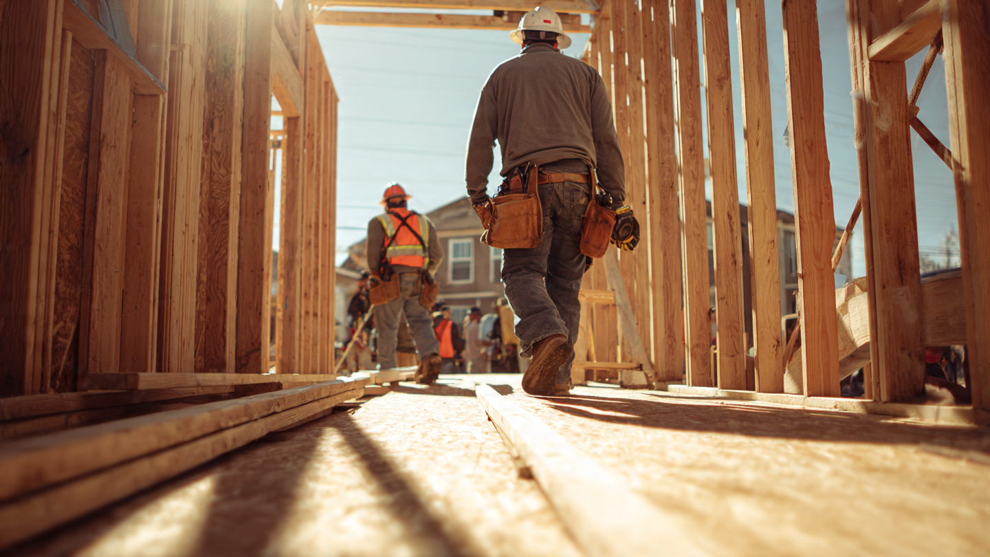 A construction worker on residential framing in midday sun, wrist bare, while a coworker nearby wears a safety monitoring band, warm harsh sunlight casting long shadows across plywood decking